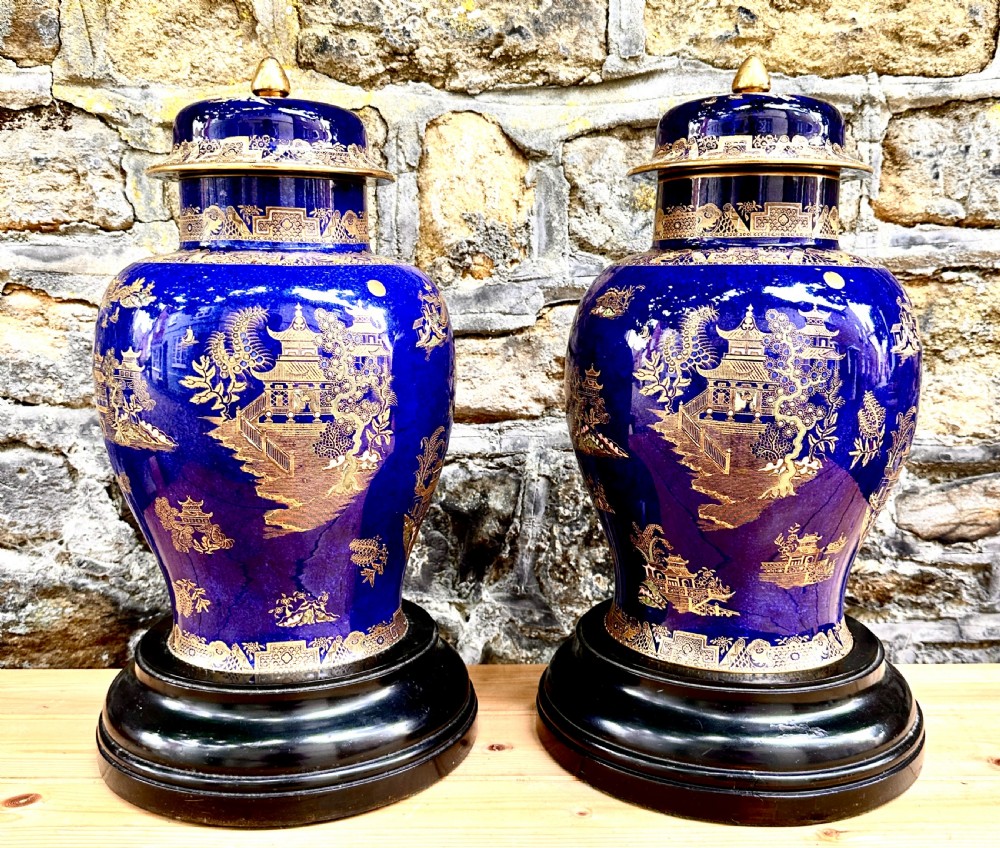 A pair of cobalt blue ceramic lidded vases decorated with gold gilt Chinoiserie pagoda scenes, set on dark circular ebonised wooden bases.