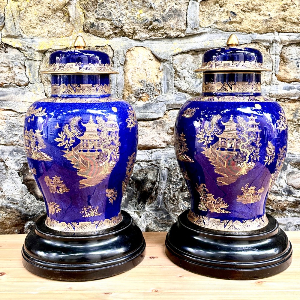 A pair of cobalt blue ceramic lidded vases decorated with gold gilt Chinoiserie pagoda scenes, set on dark circular ebonised wooden bases. View II