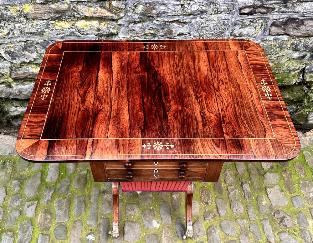 A Regency rosewood drop-leaf work table featuring wood inlay, two front drawers, a pleated pink fabric sewing basket beneath, and splayed legs with brass paw castors. View IX