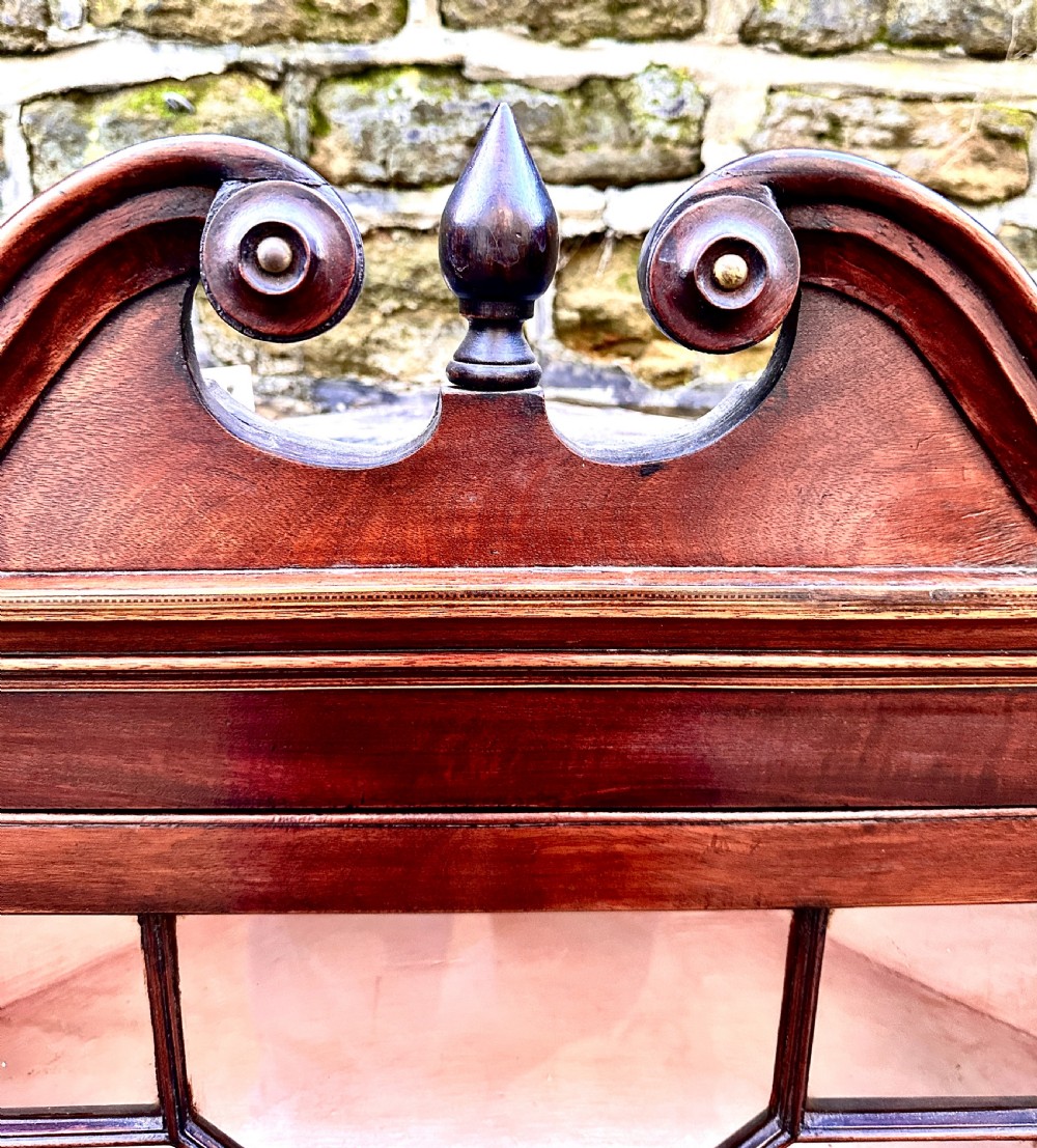 A George III mahogany corner cupboard featuring a glazed door with geometric astragal bars, a swan-neck pediment with a central finial, and brass H-hinges. View V