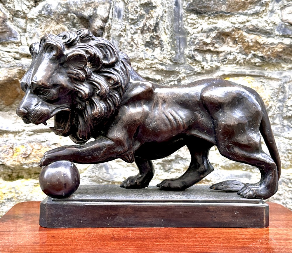 A dark brown bronze sculpture of a lion standing on a rectangular base with one paw resting upon a sphere. View II