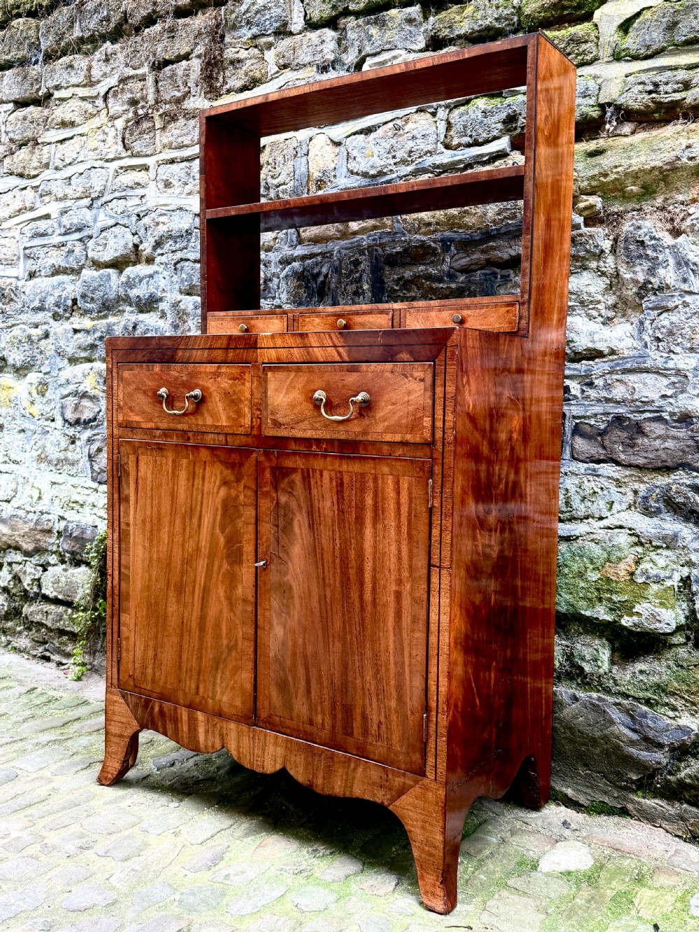 A George III mahogany chiffonier featuring a top open shelf unit with three small drawers above a two-drawer base cabinet with panelled doors and curved feet. View II