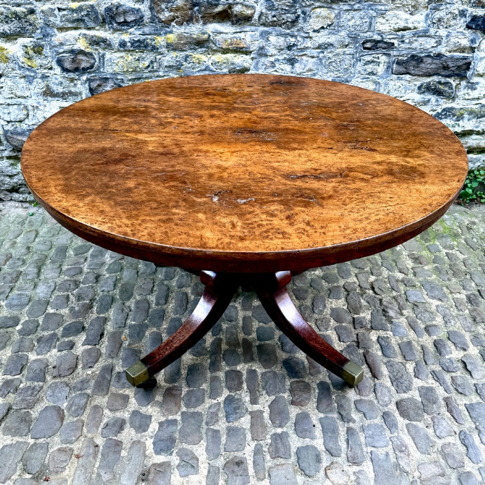 A circular George III burr yew wood pedestal centre table shown tilted vertically on downswept legs with brass castors. View VII