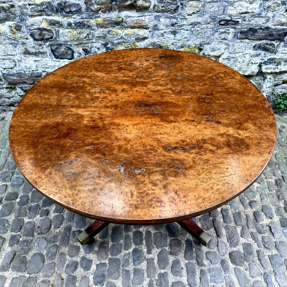 A circular George III burr yew wood pedestal centre table shown tilted vertically on downswept legs with brass castors. View VIII
