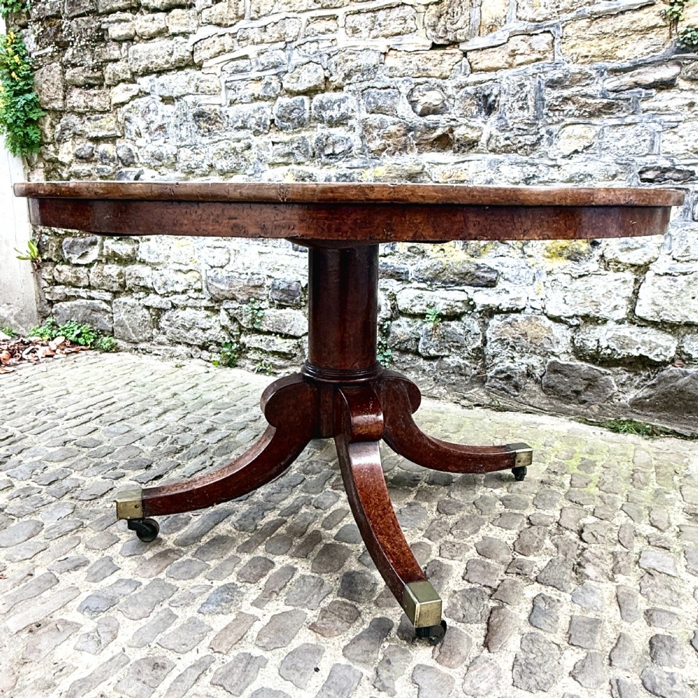 A circular George III burr yew wood pedestal centre table shown tilted vertically on downswept legs with brass castors. View IX