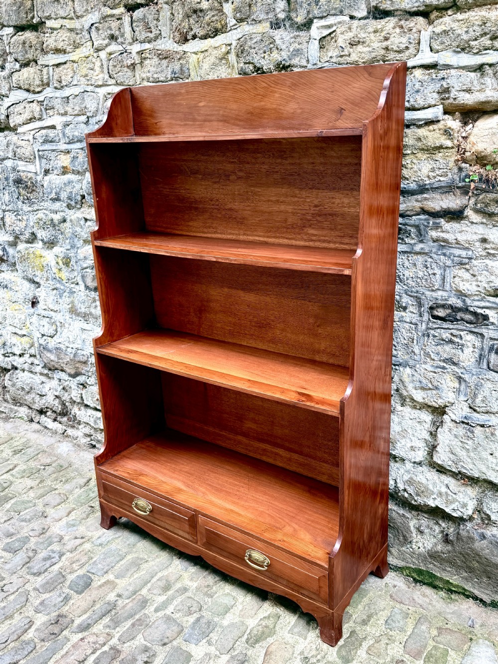 An early twentieth-century yew wood waterfall bookcase featuring four graduated open shelves above two drawers with brass handles. View II