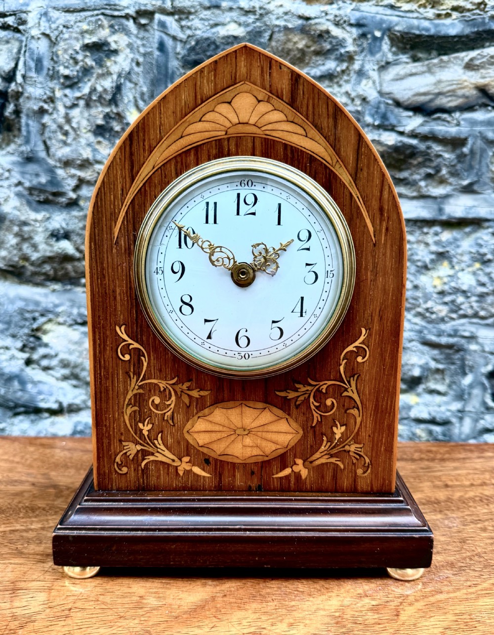 A lancet-arched wooden mantel clock featuring marquetry floral inlays, a white enamel dial, and a brass bezel.