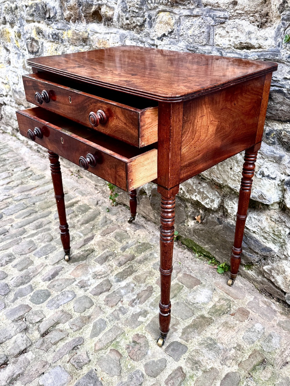 A Regency mahogany side table featuring two drawers with turned wooden handles, ring-turned legs, and brass castors. View IV