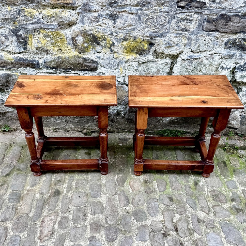 A pair of warm brown yew wood joint stools with rectangular tops, turned legs, and square perimeter stretchers resting on a cobblestone floor.