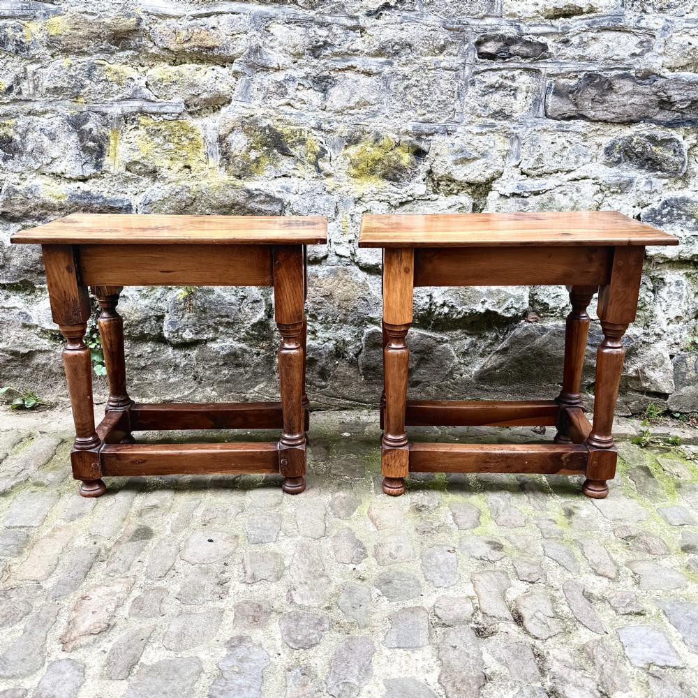 A pair of warm brown yew wood joint stools with rectangular tops, turned legs, and square perimeter stretchers resting on a cobblestone floor. View II