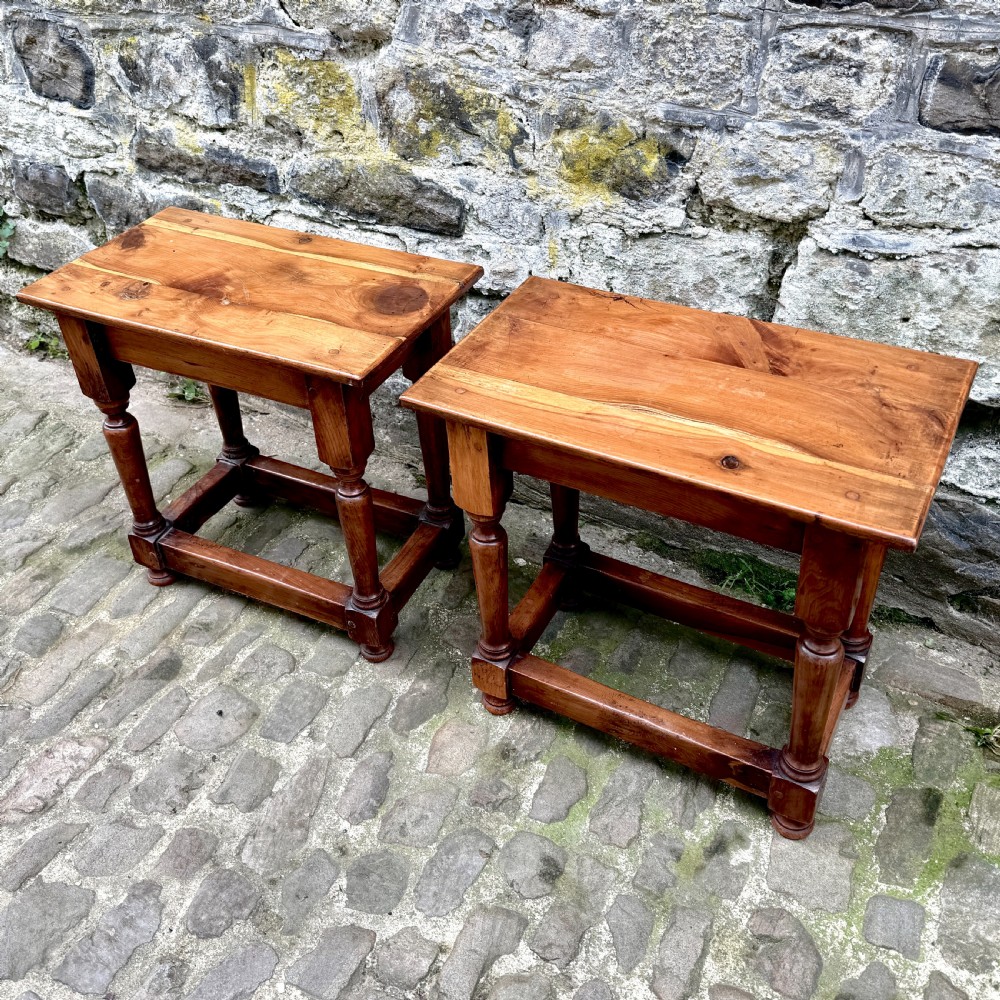 A pair of warm brown yew wood joint stools with rectangular tops, turned legs, and square perimeter stretchers resting on a cobblestone floor. View III