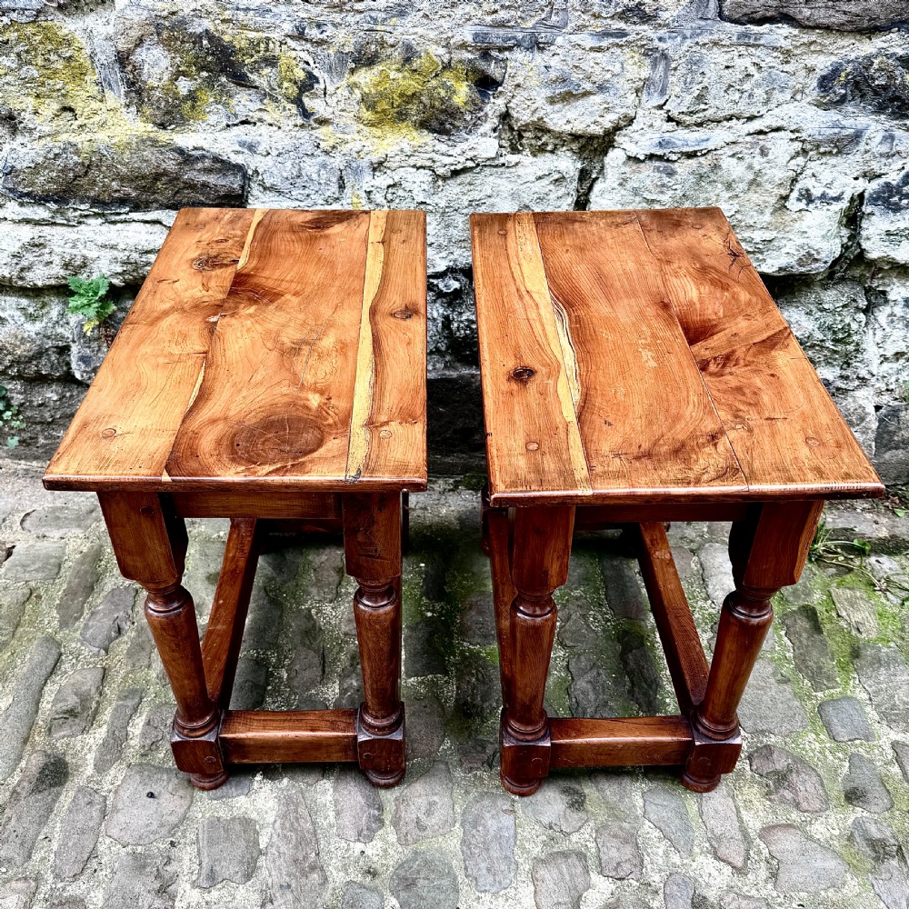 A pair of warm brown yew wood joint stools with rectangular tops, turned legs, and square perimeter stretchers resting on a cobblestone floor. View IV