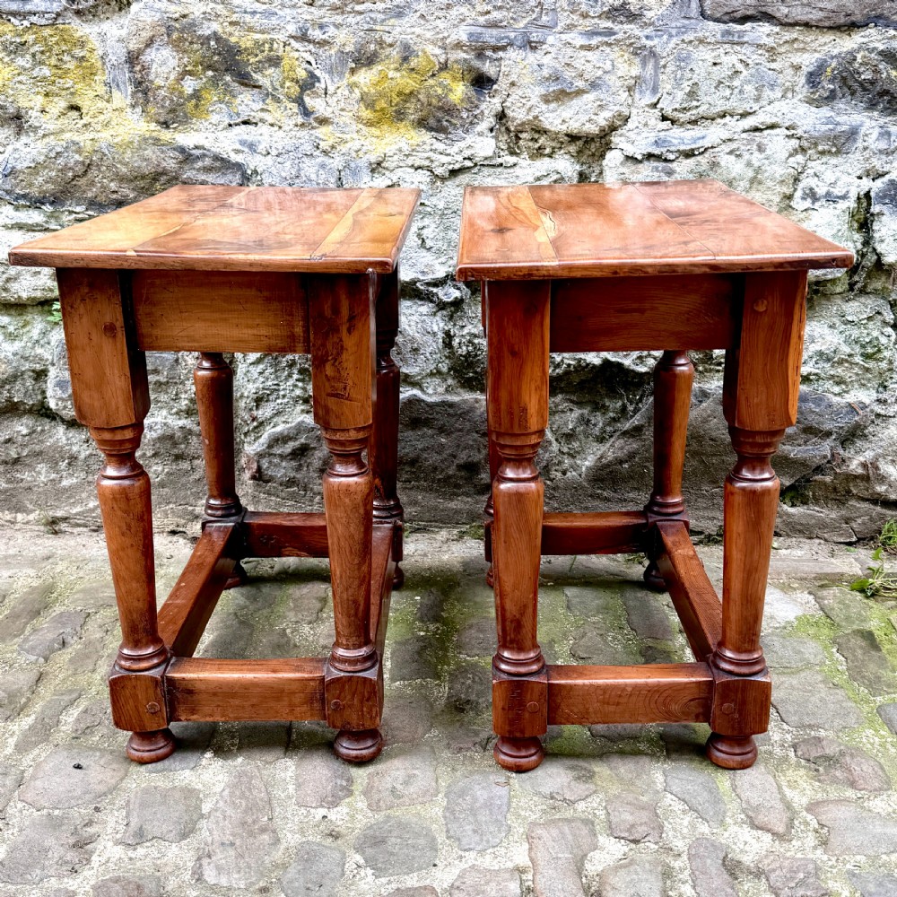 A pair of warm brown yew wood joint stools with rectangular tops, turned legs, and square perimeter stretchers resting on a cobblestone floor. View V