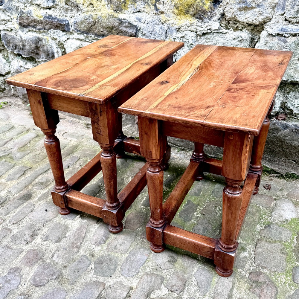 A pair of warm brown yew wood joint stools with rectangular tops, turned legs, and square perimeter stretchers resting on a cobblestone floor. View VI