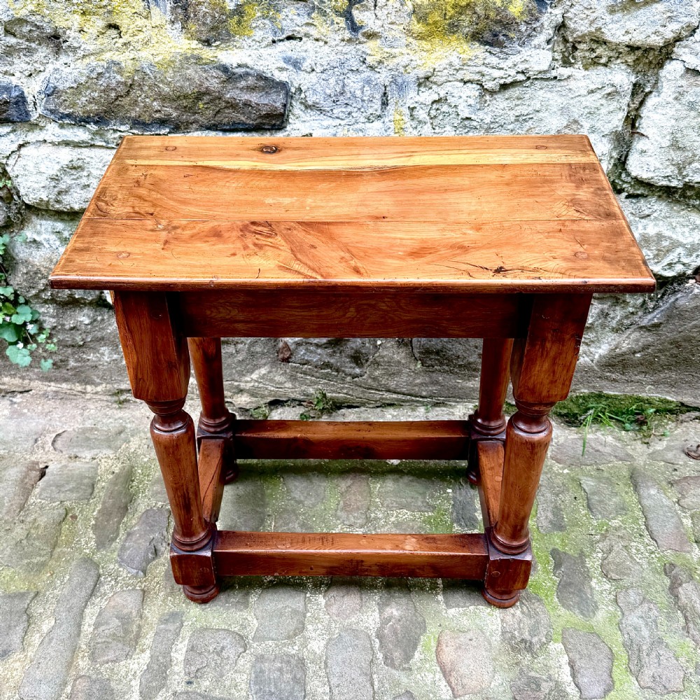 A pair of warm brown yew wood joint stools with rectangular tops, turned legs, and square perimeter stretchers resting on a cobblestone floor. View VII