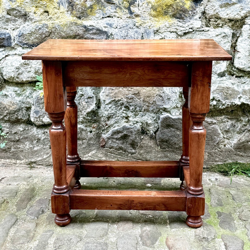 A pair of warm brown yew wood joint stools with rectangular tops, turned legs, and square perimeter stretchers resting on a cobblestone floor. View VIII