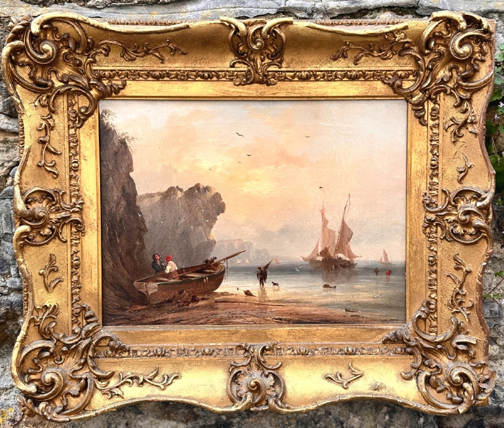 An early nineteenth-century oil painting depicting figures and boats along a rocky coastline, presented in an ornate swept giltwood frame.