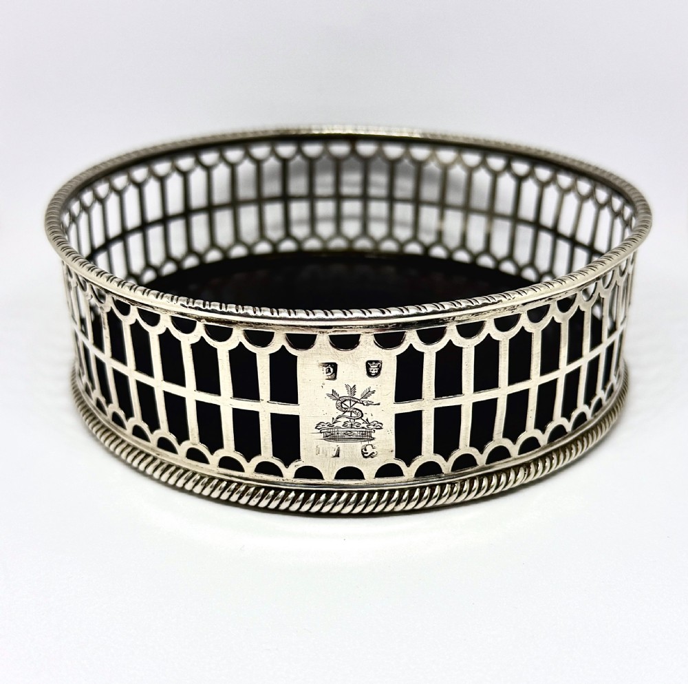 An early George III sterling silver wine coaster with pierced fretwork sides, an engraved crest, and a dark wooden base.