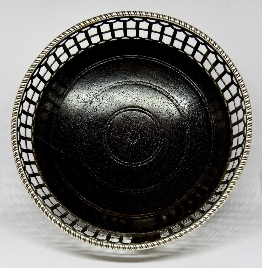 An early George III sterling silver wine coaster with pierced fretwork sides, an engraved crest, and a dark wooden base. View II