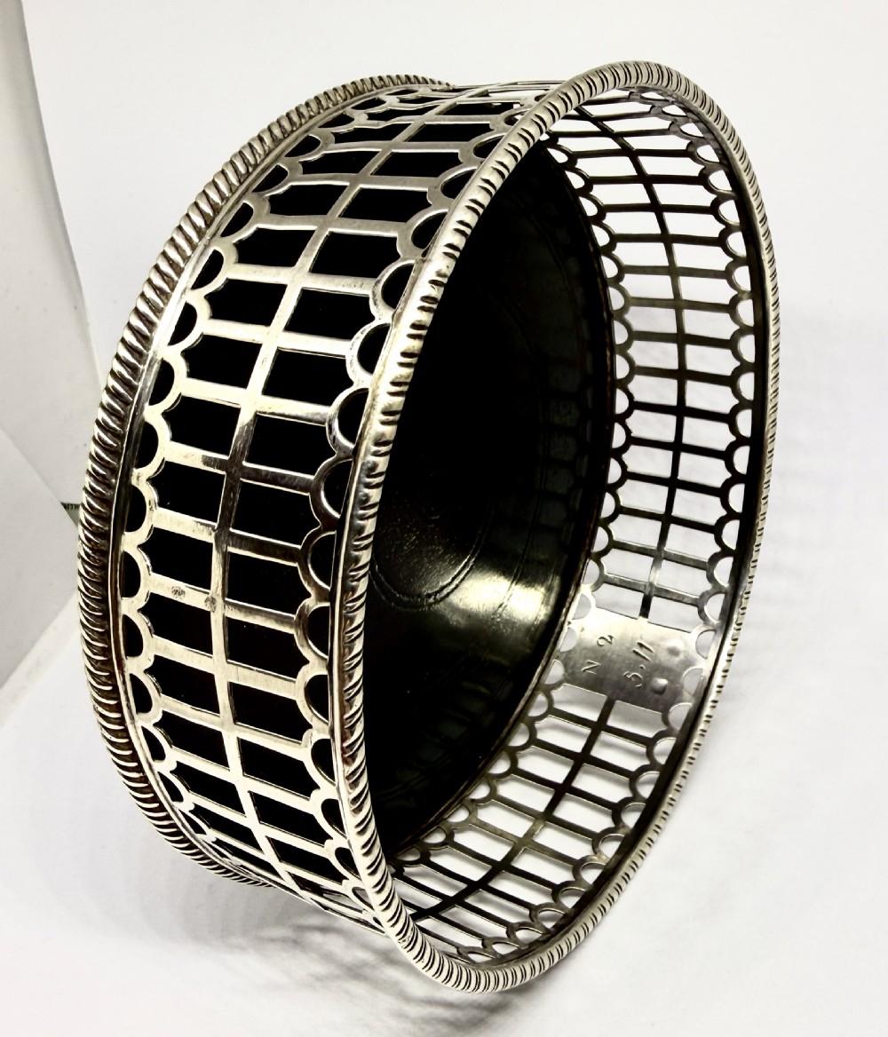 An early George III sterling silver wine coaster with pierced fretwork sides, an engraved crest, and a dark wooden base. View III