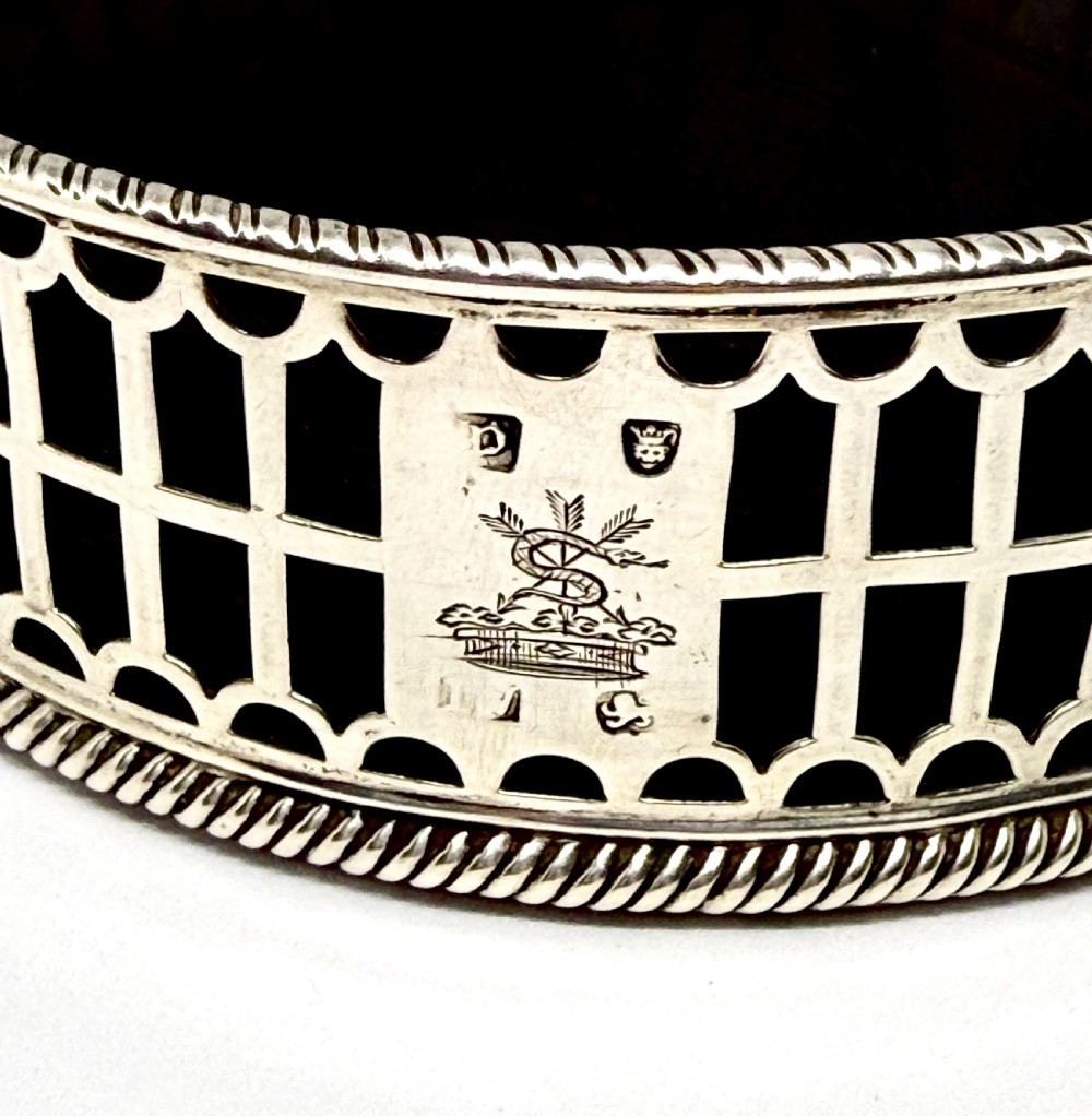 An early George III sterling silver wine coaster with pierced fretwork sides, an engraved crest, and a dark wooden base. View IV