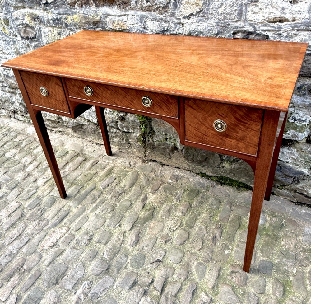 A Sheraton Revival mahogany writing table featuring a rectangular top, three drawers with brass ring drop handles, and slim tapered square legs.
