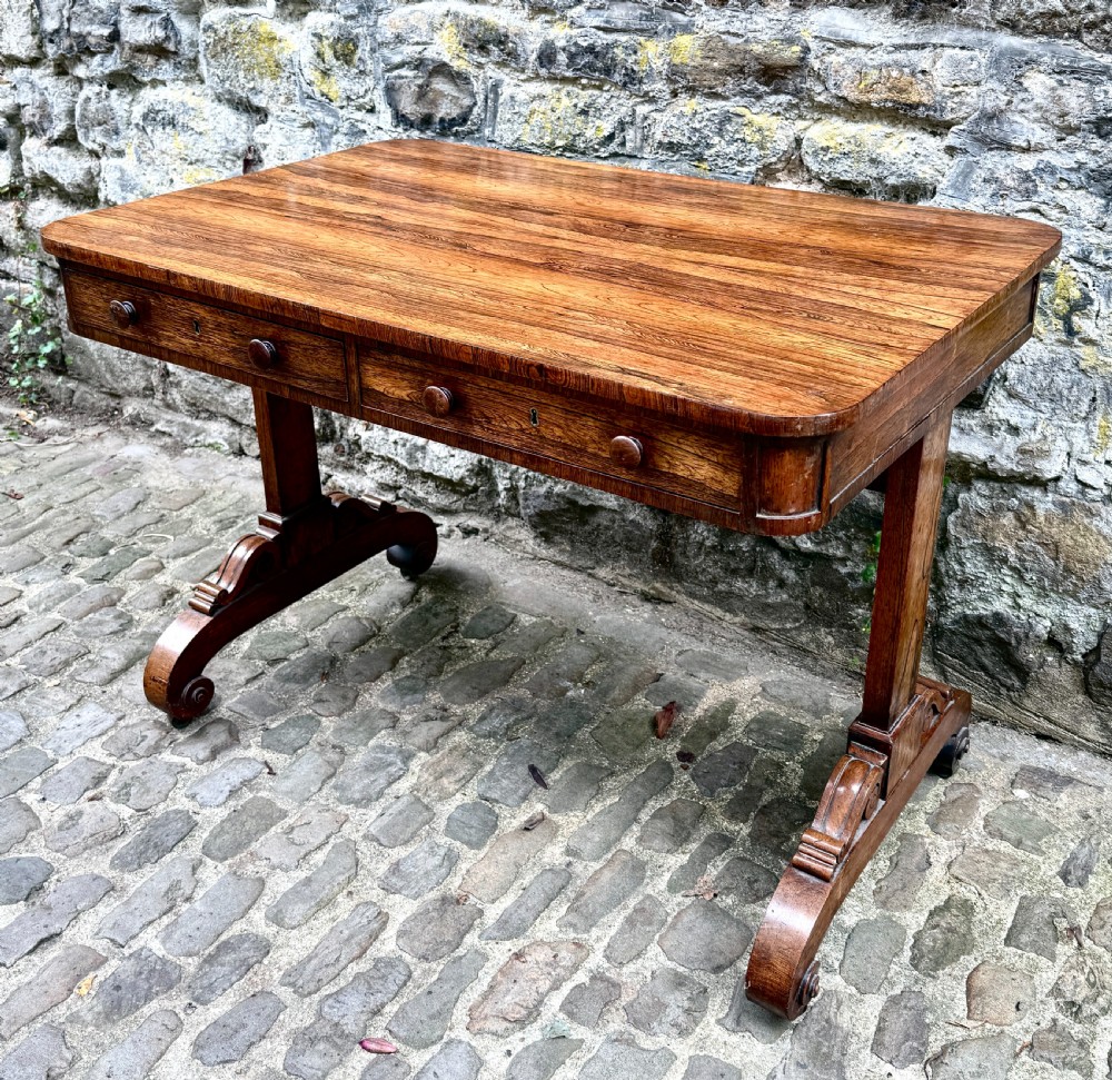 A George IV rosewood library table with a figured timber top, frieze drawers, turned handles, and carved scrolled feet.