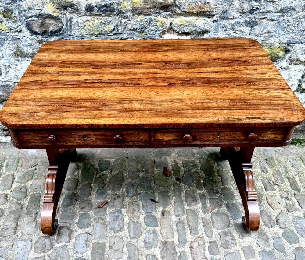 A George IV rosewood library table with a figured timber top, frieze drawers, turned handles, and carved scrolled feet. View II