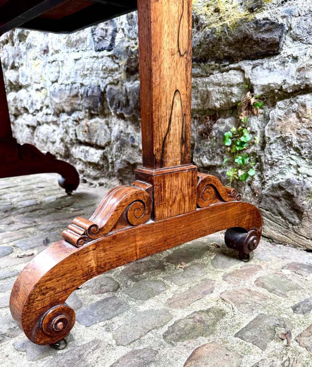 A George IV rosewood library table with a figured timber top, frieze drawers, turned handles, and carved scrolled feet. View IV