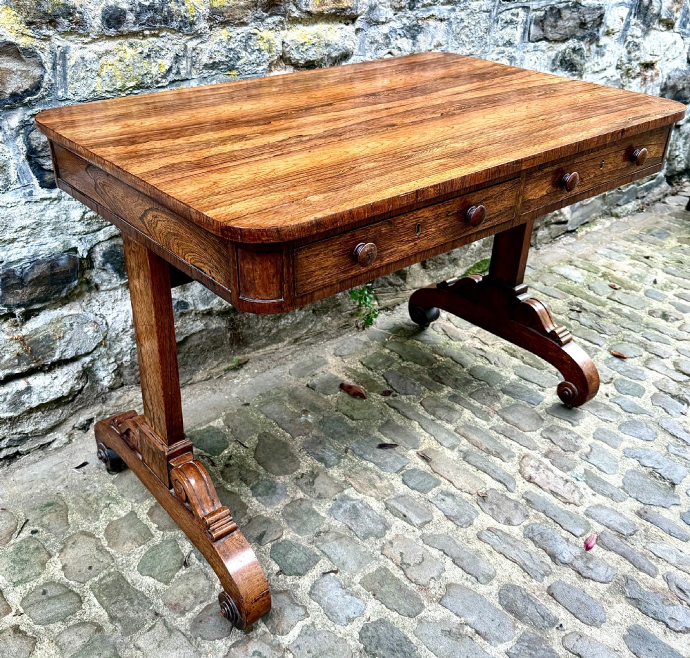 A George IV rosewood library table with a figured timber top, frieze drawers, turned handles, and carved scrolled feet. View VII