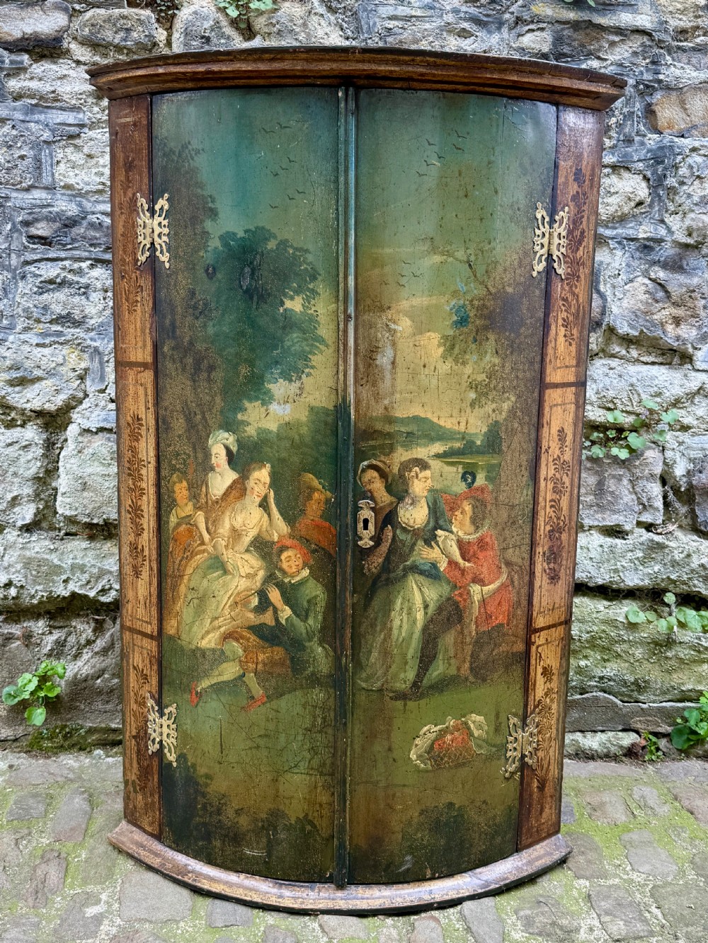 An eighteenth-century George II bowfront corner cupboard featuring two curved doors painted with a green pastoral scene and fitted with pierced brass hinges.
