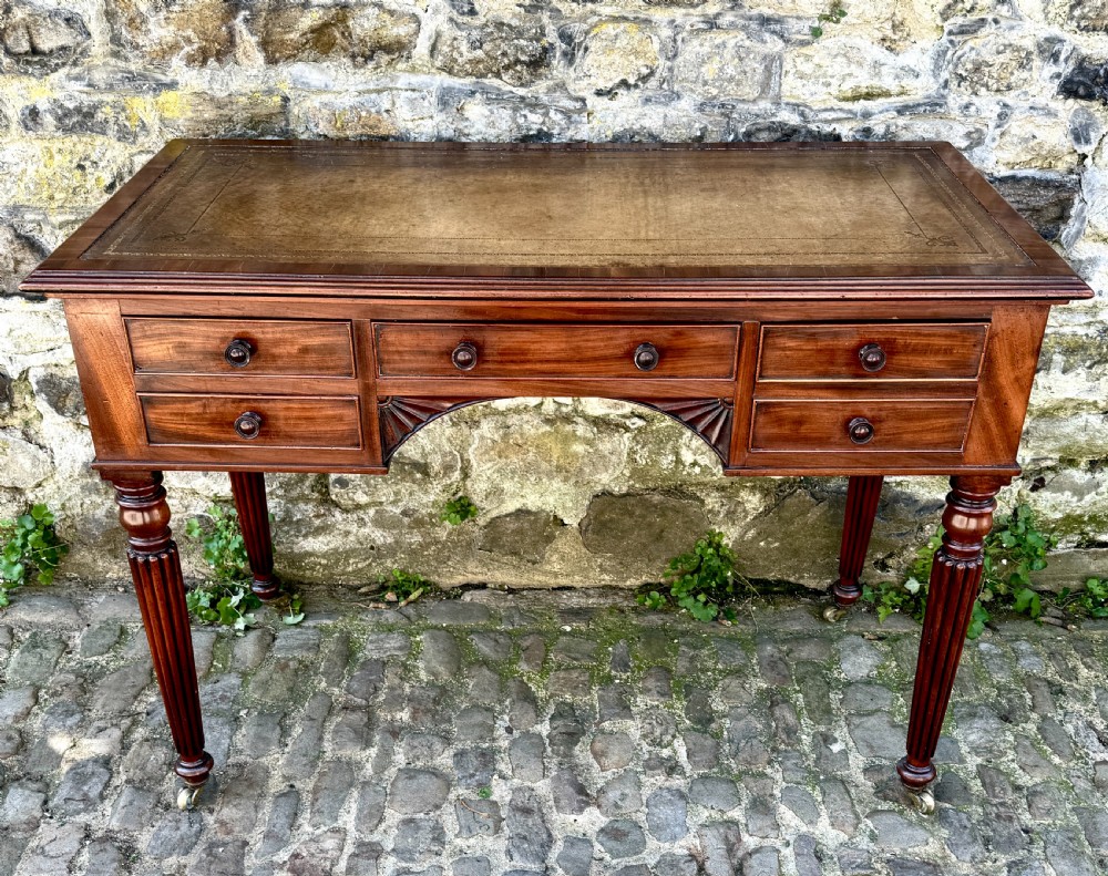 A George IV mahogany writing table featuring a gold-tooled brown leather top, five drawers, carved fan spandrels, and reeded legs on brass castors.