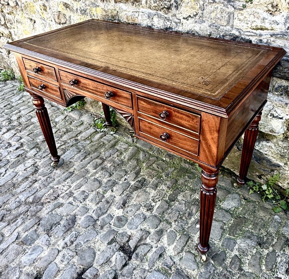 A George IV mahogany writing table featuring a gold-tooled brown leather top, five drawers, carved fan spandrels, and reeded legs on brass castors. View II