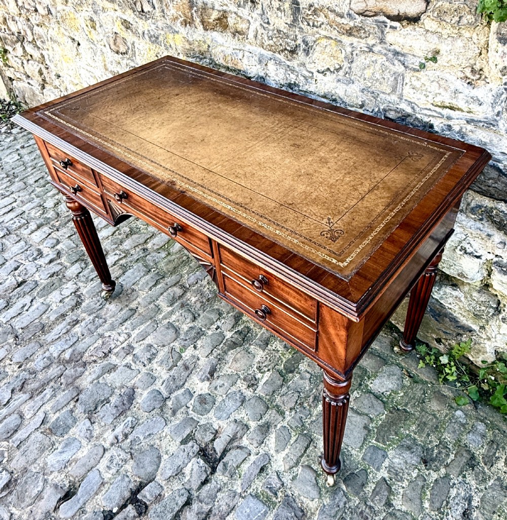 A George IV mahogany writing table featuring a gold-tooled brown leather top, five drawers, carved fan spandrels, and reeded legs on brass castors. View III