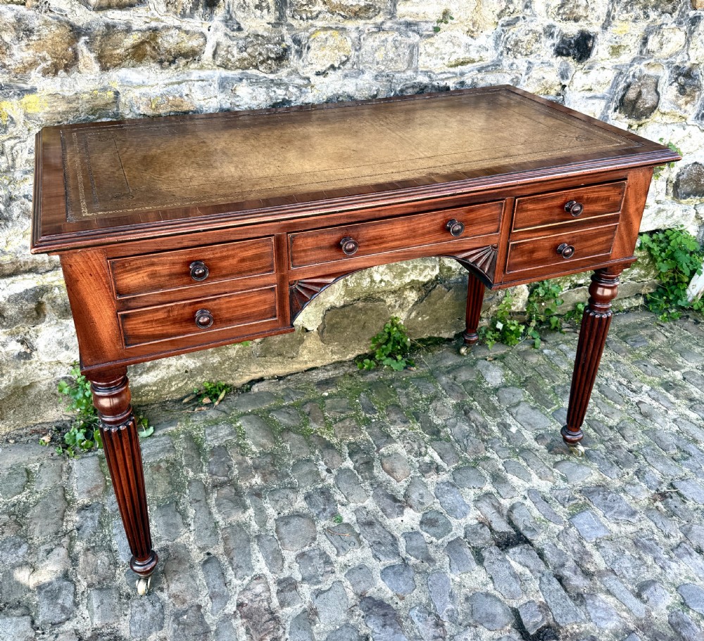 A George IV mahogany writing table featuring a gold-tooled brown leather top, five drawers, carved fan spandrels, and reeded legs on brass castors. View IV