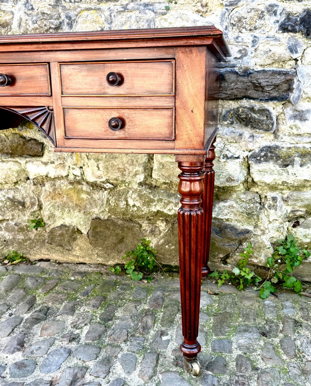 A George IV mahogany writing table featuring a gold-tooled brown leather top, five drawers, carved fan spandrels, and reeded legs on brass castors. View V