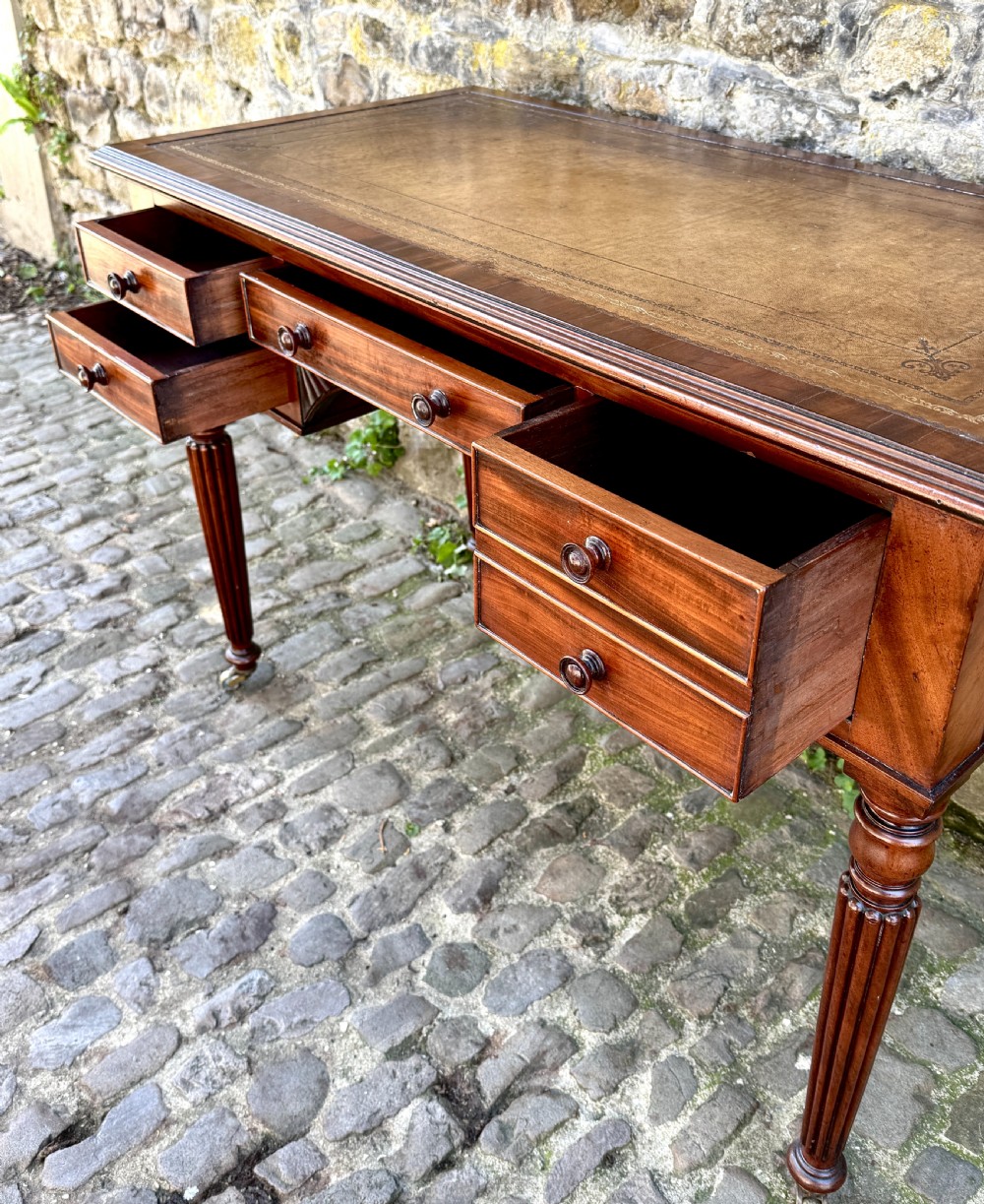 A George IV mahogany writing table featuring a gold-tooled brown leather top, five drawers, carved fan spandrels, and reeded legs on brass castors. View VII