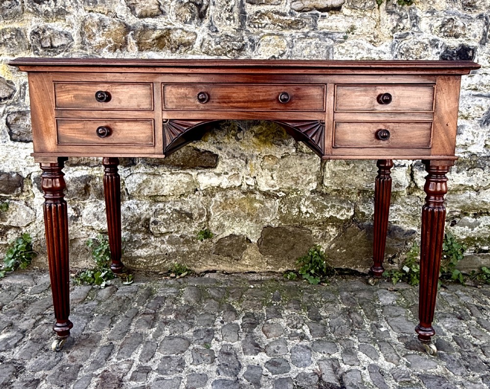 A George IV mahogany writing table featuring a gold-tooled brown leather top, five drawers, carved fan spandrels, and reeded legs on brass castors. View VIII