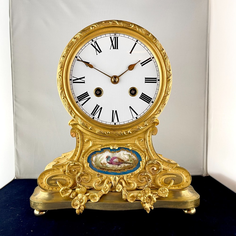 A late 19th-century French gilt bronze drumhead mantel clock featuring a white enamel dial with Roman numerals, ornate floral swags, and an oval painted porcelain plaque depicting a bird.
