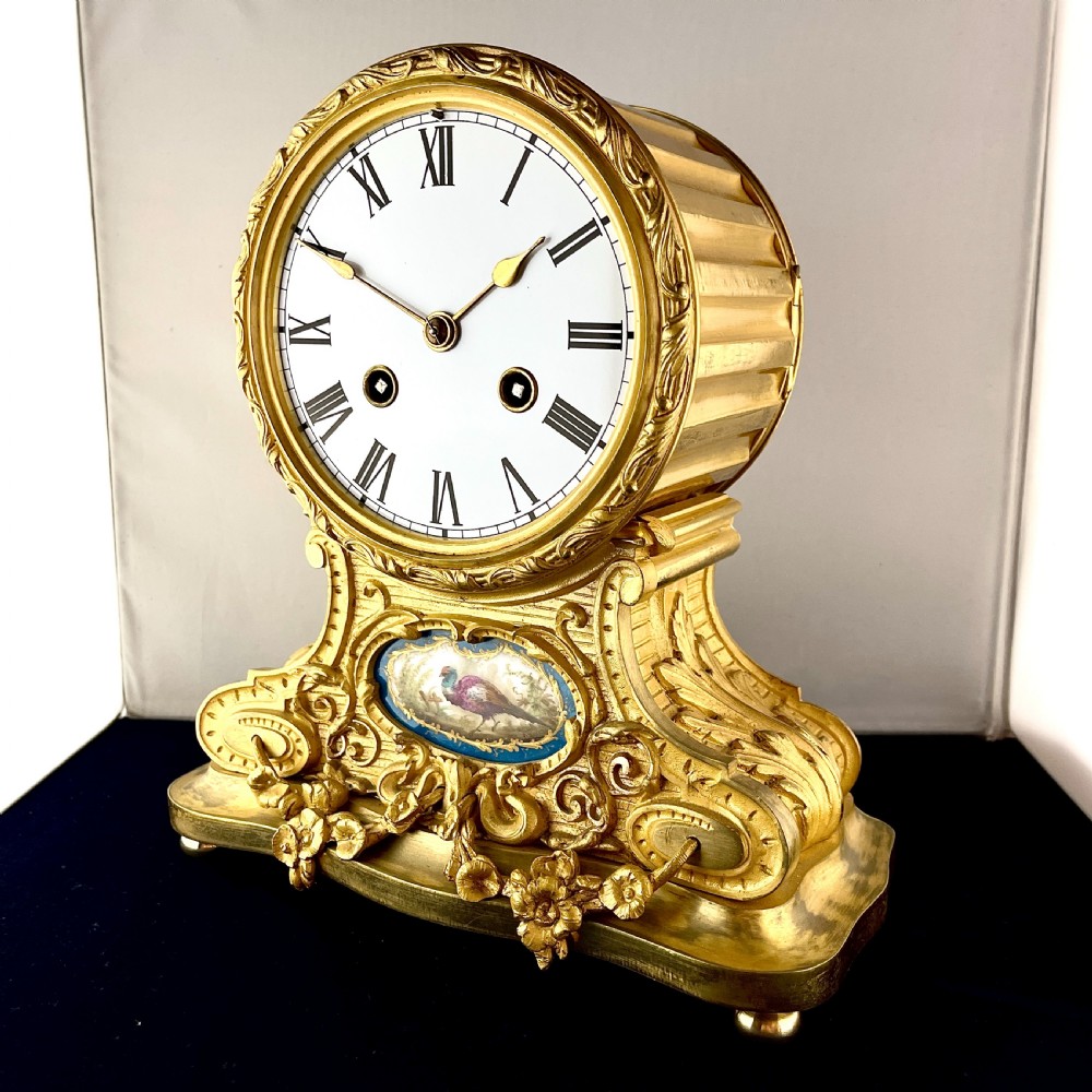 A late 19th-century French gilt bronze drumhead mantel clock featuring a white enamel dial with Roman numerals, ornate floral swags, and an oval painted porcelain plaque depicting a bird. View II