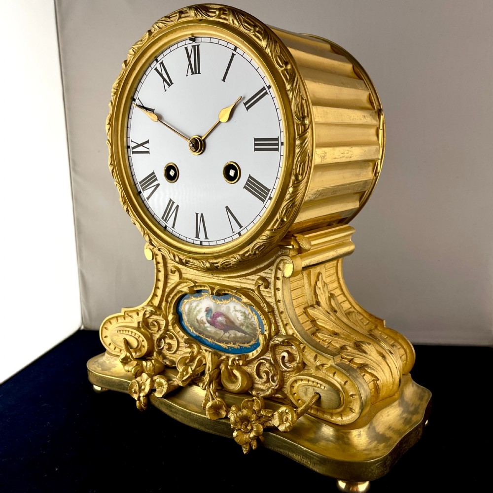 A late 19th-century French gilt bronze drumhead mantel clock featuring a white enamel dial with Roman numerals, ornate floral swags, and an oval painted porcelain plaque depicting a bird. View III