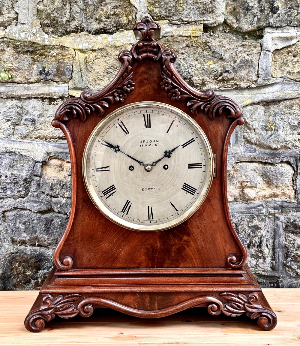 A mid-Victorian mahogany bracket clock featuring a carved scroll case, silvered circular dial with Roman numerals, and an ornate plinth base.
