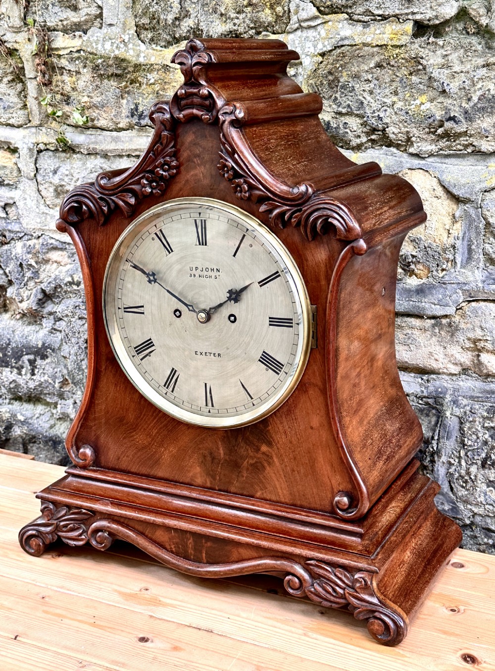 A mid-Victorian mahogany bracket clock featuring a carved scroll case, silvered circular dial with Roman numerals, and an ornate plinth base. View II