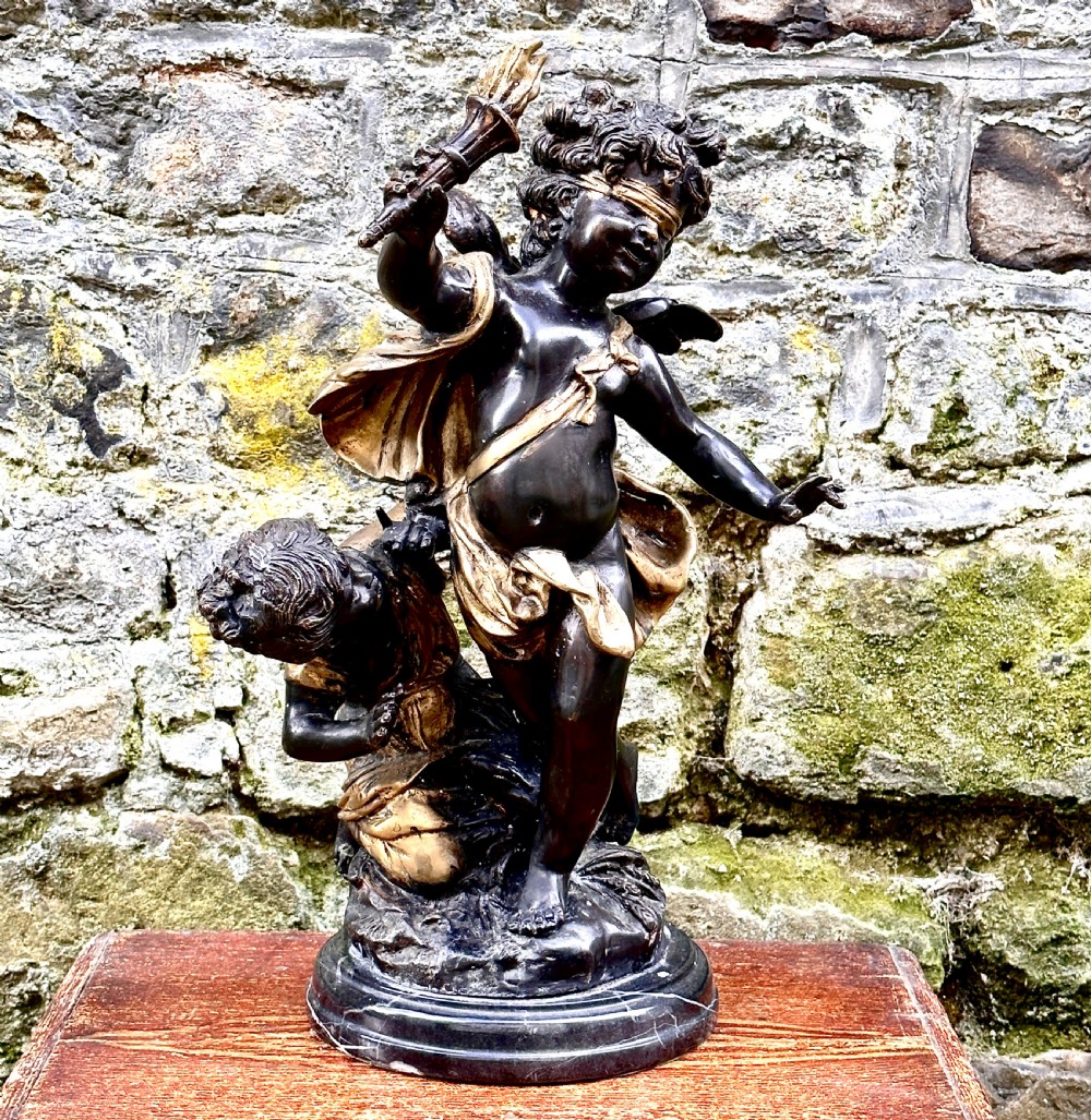 A dark bronze and parcel-gilt French sculpture depicting a blindfolded winged cherub holding a flaming torch, mounted on a round black marble base.