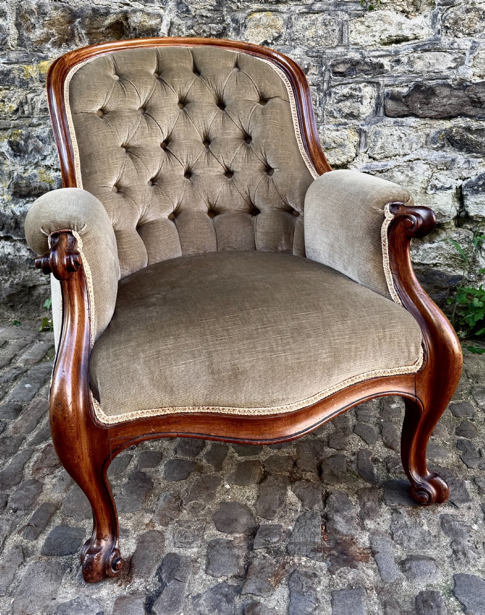 An Early Victorian walnut armchair featuring deep button-tufted olive green velvet upholstery, carved padded armrests, and cabriole front legs.