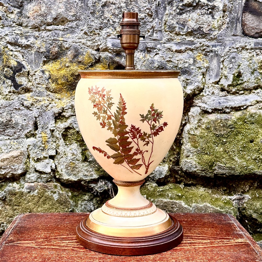 An urn-shaped cream porcelain table lamp decorated with painted fern foliage, mounted on a turned wooden base with a brass socket.