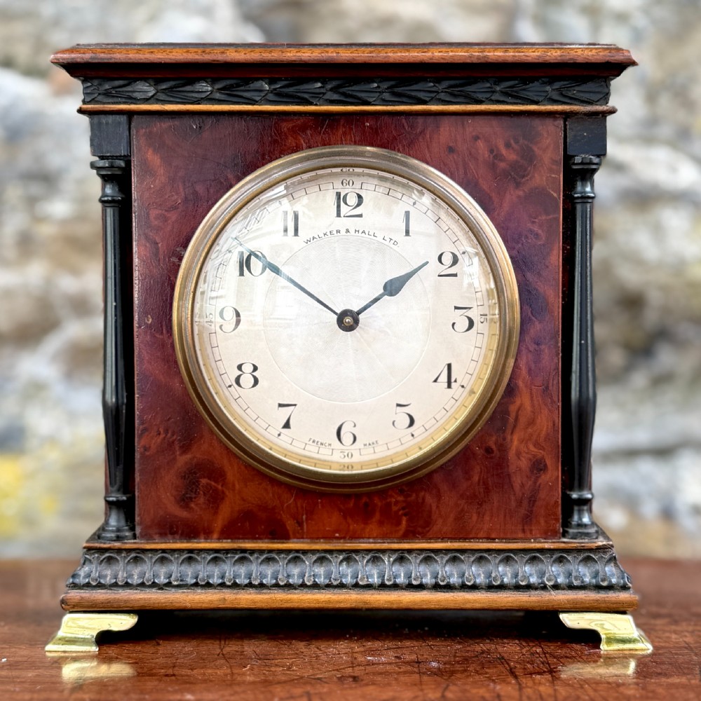 An early 20th-century rectangular mahogany and burr walnut mantel clock featuring ebonised side columns, a white circular dial signed Walker & Hall Ltd., and brass bracket feet.