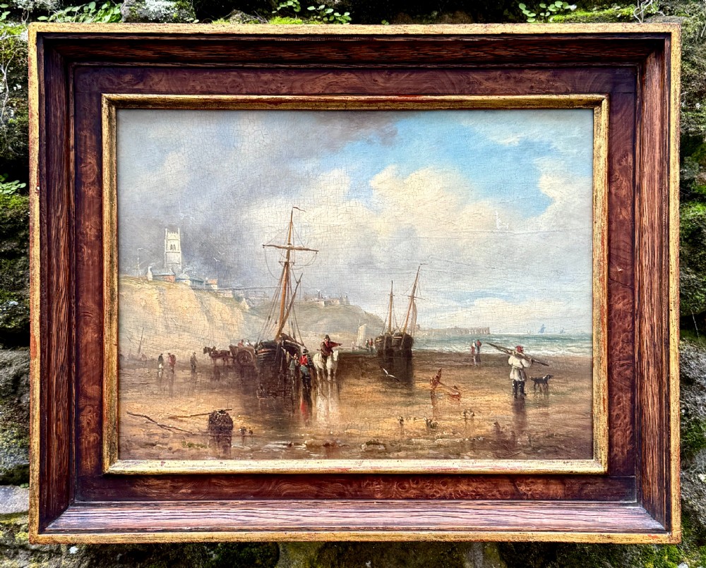 A 19th-century oil painting depicting a coastal seascape with figures, horses, and boats on a shoreline below cliffs, set within a wide timber frame with a gilded inner slip.