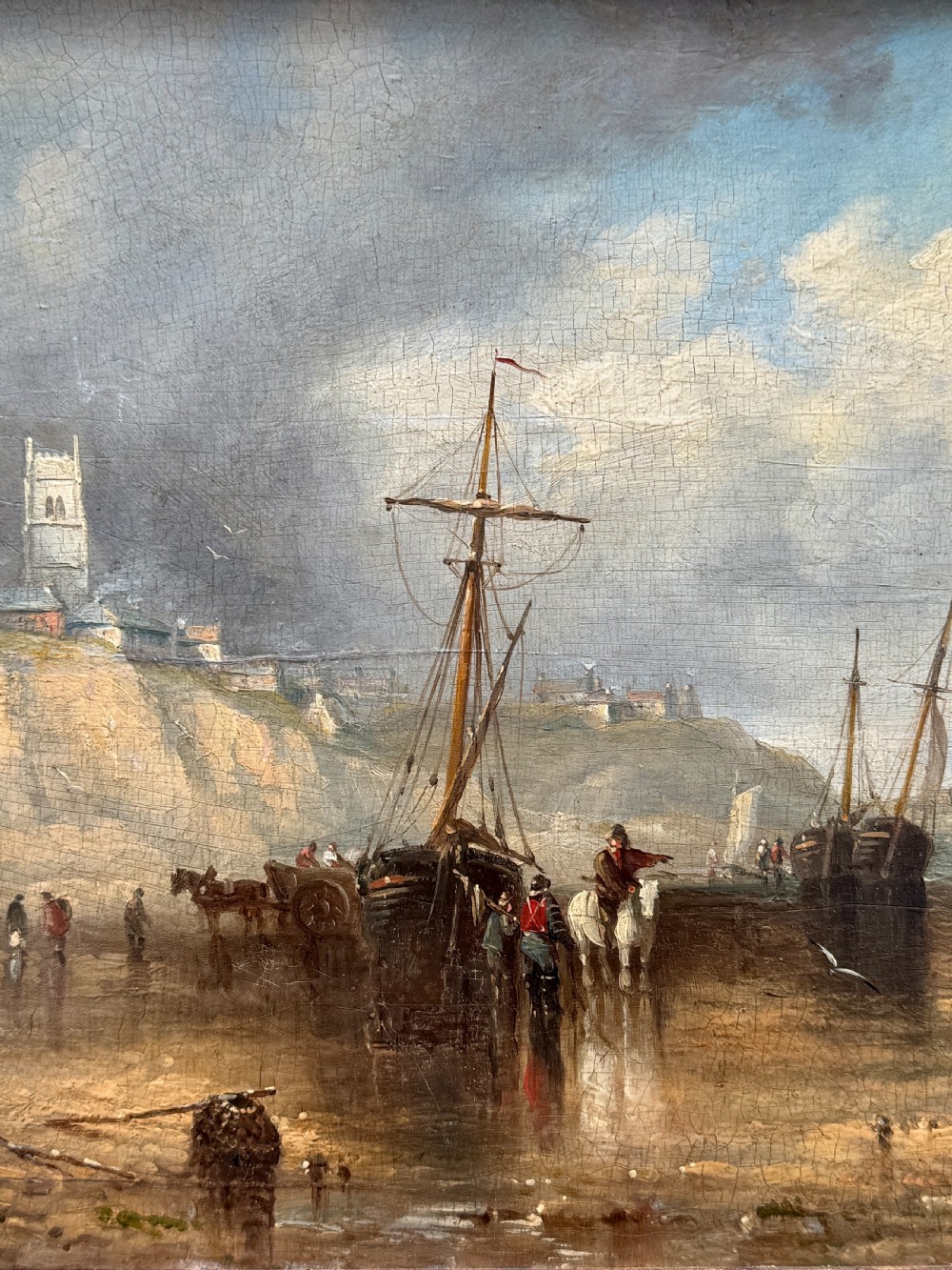 A 19th-century oil painting depicting a coastal seascape with figures, horses, and boats on a shoreline below cliffs, set within a wide timber frame with a gilded inner slip. View III