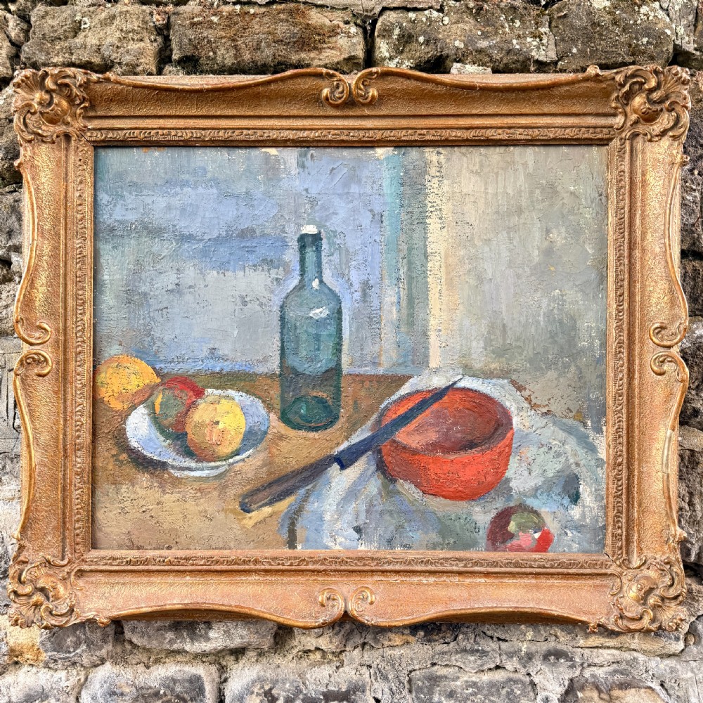 An early twentieth-century post-impressionist oil painting depicting a tabletop still life with a glass bottle, fruit, and a red bowl, enclosed within a carved swept gilt frame.